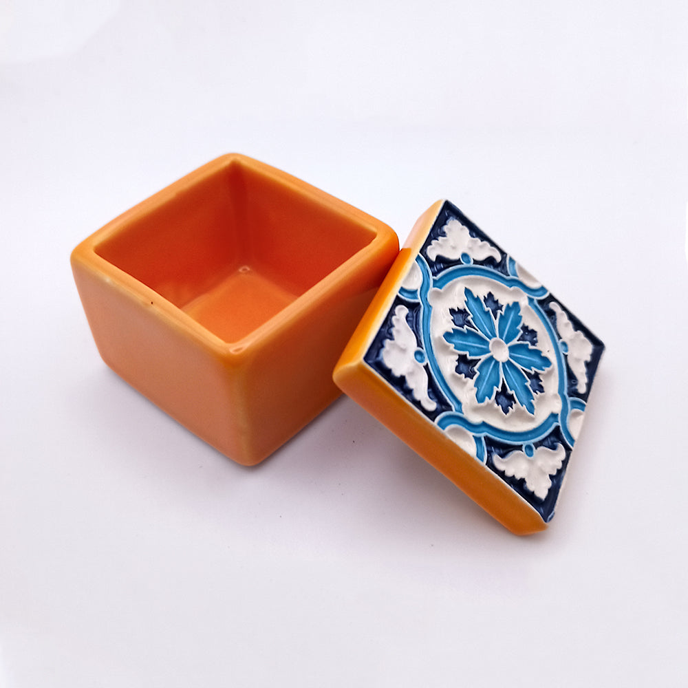 A Saint Maria Island Ceramic Box, small and square in shape, sits with its lid resting beside it. This charming orange piece features an exquisite blue and white floral pattern on the lid, making it a luxurious addition to any space.