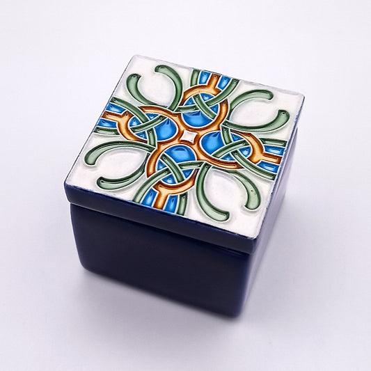 The Santarém Ceramic Box by Tejo Shop is a small, square ceramic masterpiece with a dark blue base. Its lid showcases exquisite artistry, featuring an intricate multicolored design in green, blue, and orange against a white background, reminiscent of geometric or Celtic knot patterns. The box rests on a plain white surface.