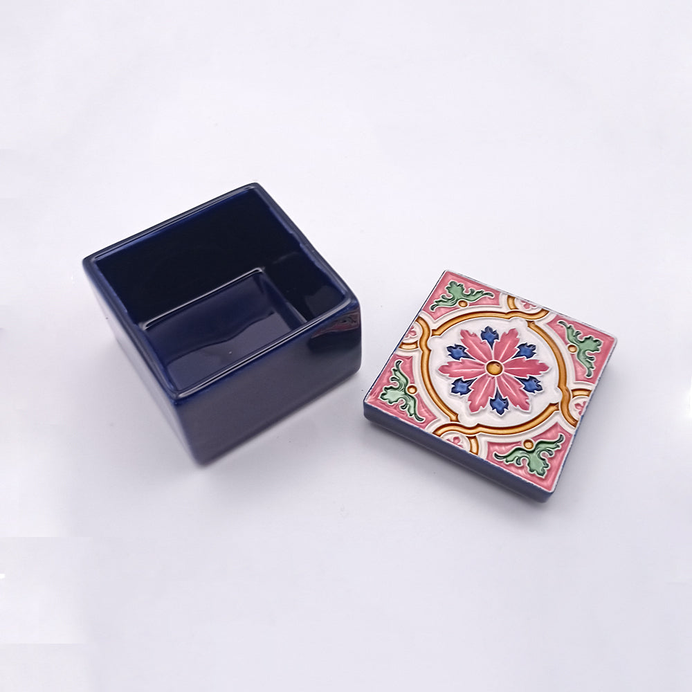 A small, square, dark blue handcrafted Tejo Shop Saint Jorge Island Ceramic Box with a removable lid. The lid boasts an exquisite colorful pattern featuring floral and geometric designs in pink, blue, and yellow hues. Both items are placed on a plain white background.