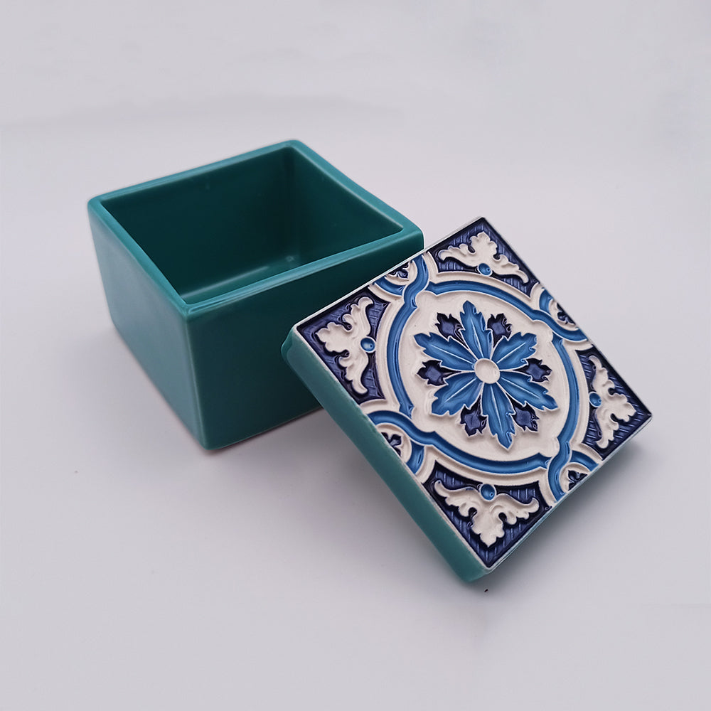 A small handcrafted square ceramic box with its lid resting beside it. The São Miguel Island Ceramic Box by Tejo Shop is turquoise, and the lid features an ornate blue and white floral pattern. The interior of the box is visible, showcasing its turquoise color. The background is a plain, soft white. A beautiful addition to any space.