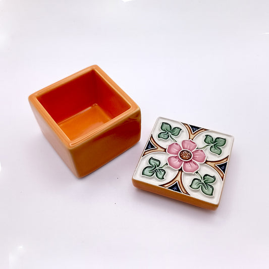 The Sintra Ceramic Box by Tejo Shop is a small square ceramic piece with an orange base and a detachable lid. The lid features a colorful floral design with a pink flower at the center, surrounded by green leaves and a decorative pattern, exuding handcrafted artistic charm. The box is open, showing its empty interior.