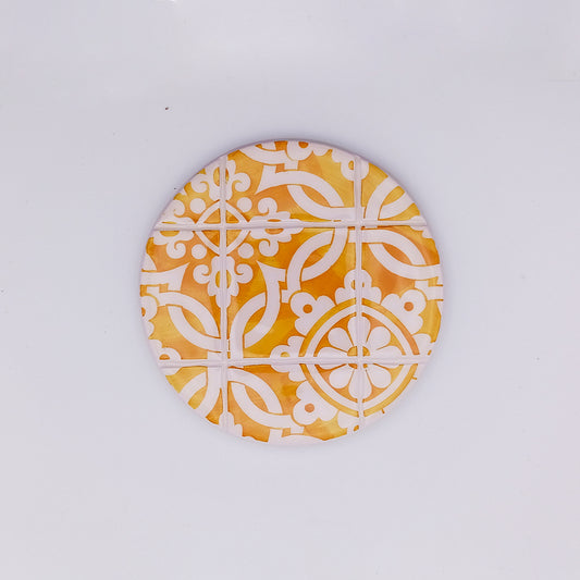 A round, hand-made coaster featuring a geometric pattern in white and varying shades of yellow and orange. The design, reminiscent of a traditional Sintra Ceramic Cup Pads by Tejo Shop, consists of symmetrically arranged curved and angular shapes on a plain white background.