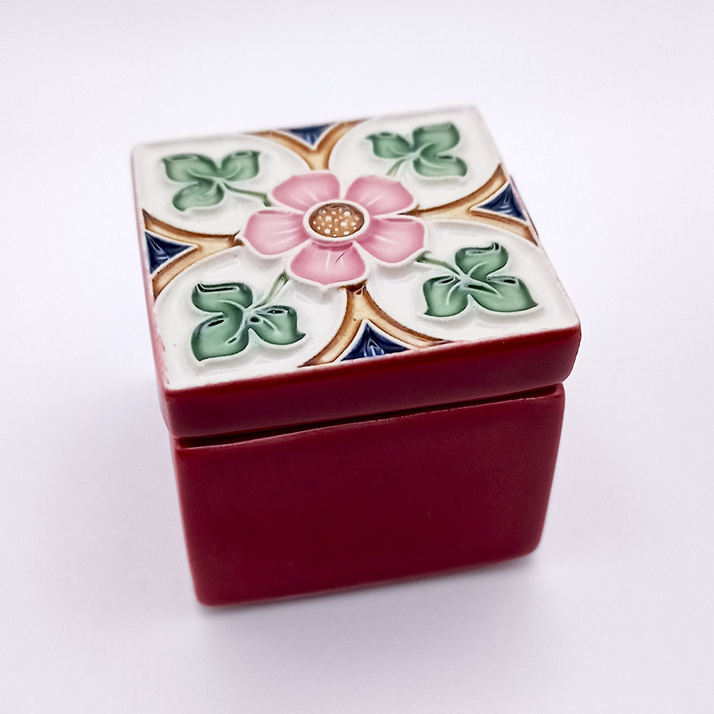 A small, square red ceramic box with a decorative lid featuring a raised floral design. Handcrafted in the style of Tavira Ceramic Box by Tejo Shop, the lid has a central pink flower surrounded by green leaves and blue and gold accents, set against a white background. The slightly ajar lid reveals the box’s interior.