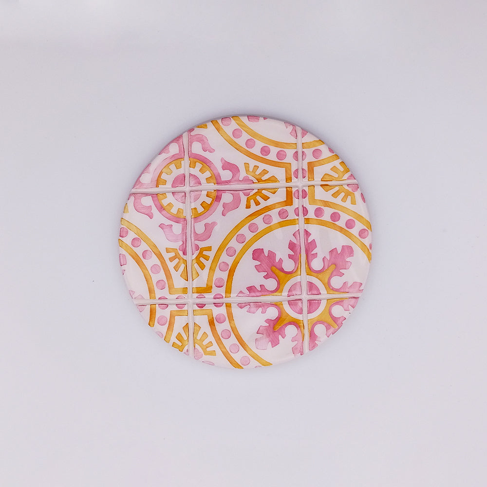 A round ceramic tile coaster, perfect for home decor, featuring a decorative pattern with pink and yellow geometric and floral designs. The symmetrical, hand-painted design has curving lines and intricate accents that form a colorful motif on a white background. Ideal as the Tejo Shop Tavira Ceramic Cup Pad.