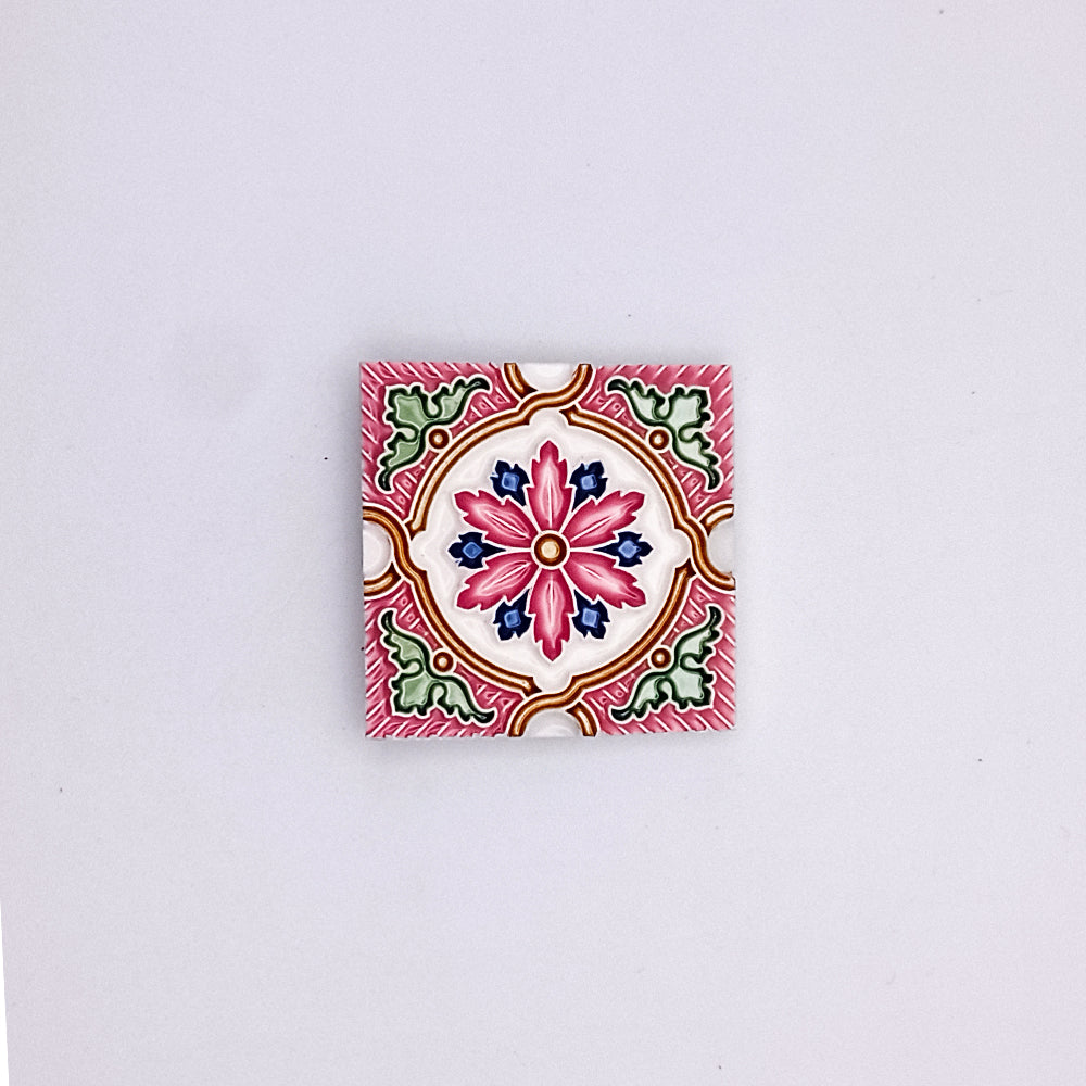 A vibrant, square ceramic tile with a detailed Small Spanish Tile Pattern in pink, red, green, and white, centered on a light gray background by Tejo Shop.