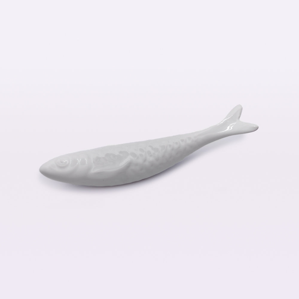 A handcrafted white ceramic object shaped like a fish, with detailed scales and fins, lies horizontally on a plain, light-colored surface. This exquisite piece of Portuguese Ceramic Sardines by Tejo Shop makes an elegant addition to any home decor.